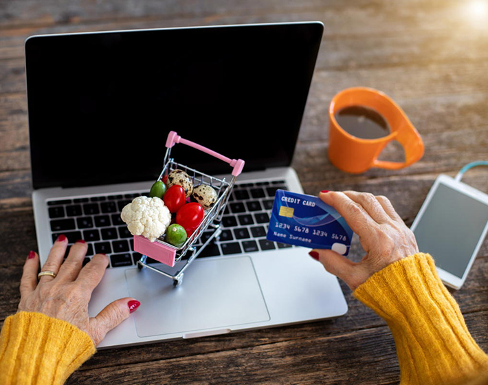 senior woman at a laptop with a credit card and tiny shopping cart to suggest online shopping and delivery