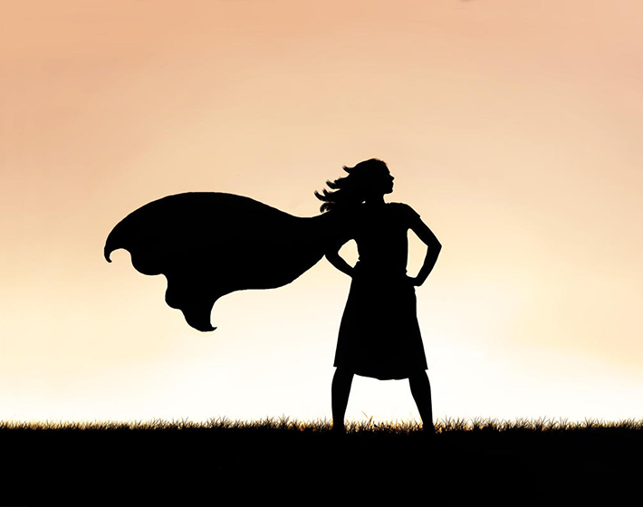 silhouette of a woman in a strong pose wearing a superhero cape against a sunset