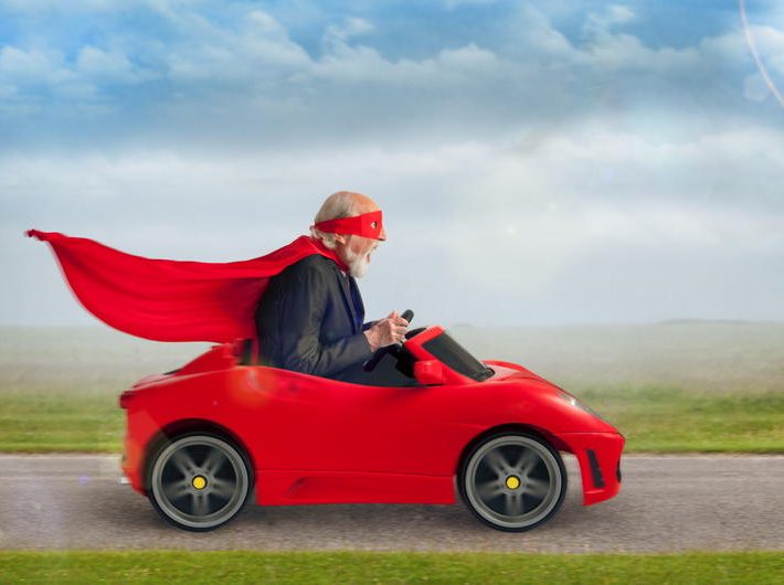 senior superhero with mask and cape driving a toy sports car